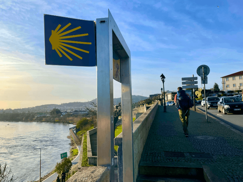 A pilgrim walking next to a Camino de Santiago scallop shell marker next to the C&aacute;vado River in Barcelos, Portugal