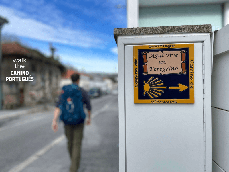 A pilgrim walking along the street next to a sign that reads "Aqui vive en peregrino" (here lives a pilgrim)